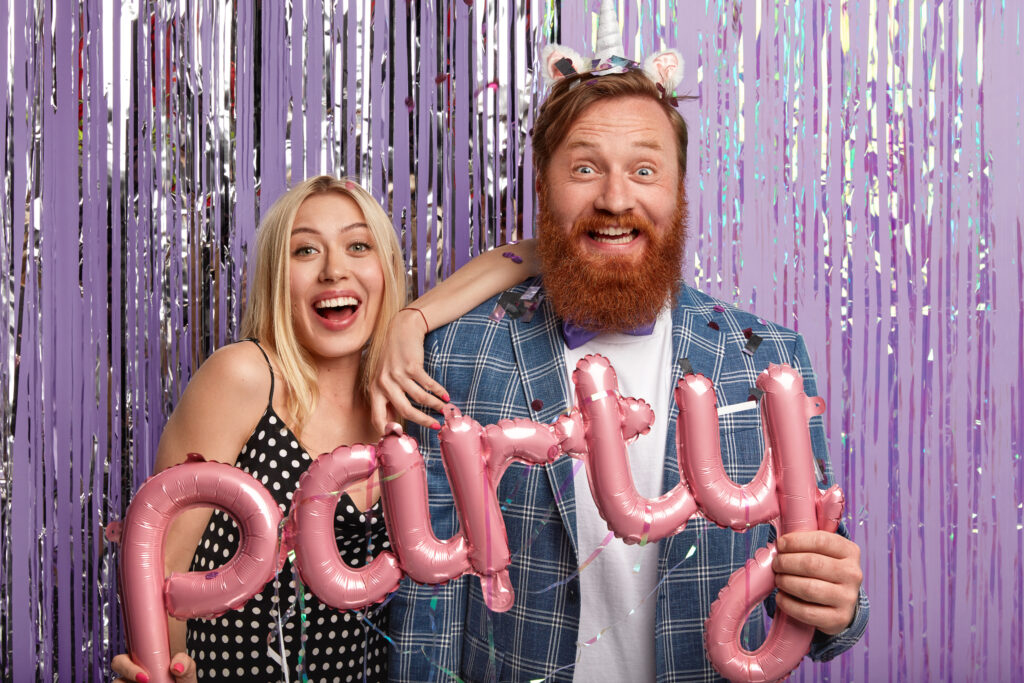 Joyous couple stand next to each other, have fun indoor, pose in decorated photozone, enjoys spare time, hold pink balloons in form of letters, have overjoyed facial expressions invite friends to join svg+xml;charset=utf